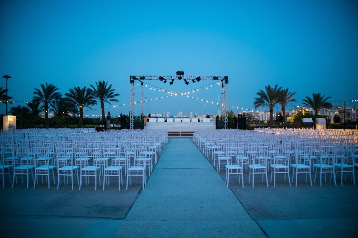 An empty outdoor event lawn at blue hour, with rows of white chairs either side of a wide central aisle, a lighting truss over a low stage, string lights and palm trees, and a city skyline in the distance.